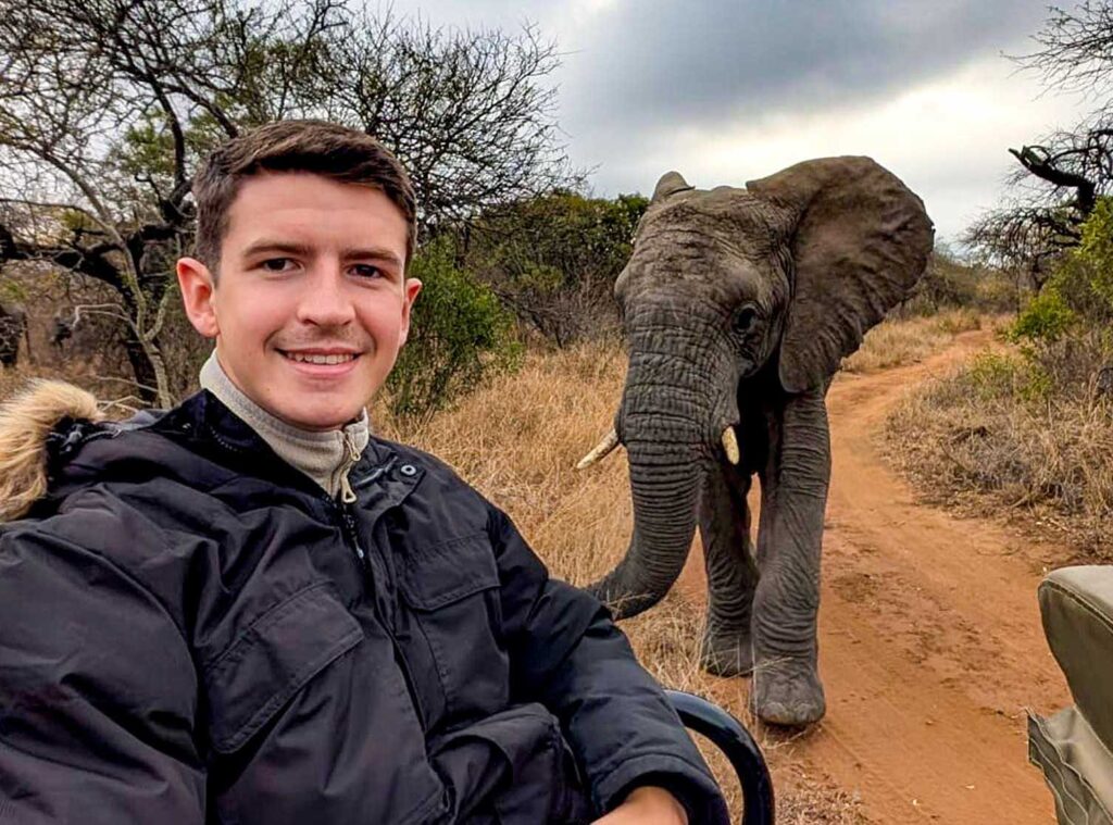 A picture of a young man smiling on a safari jeep with a camera on his lap, with a young African Elephant a few metres behind on a dirt track in South Africa.